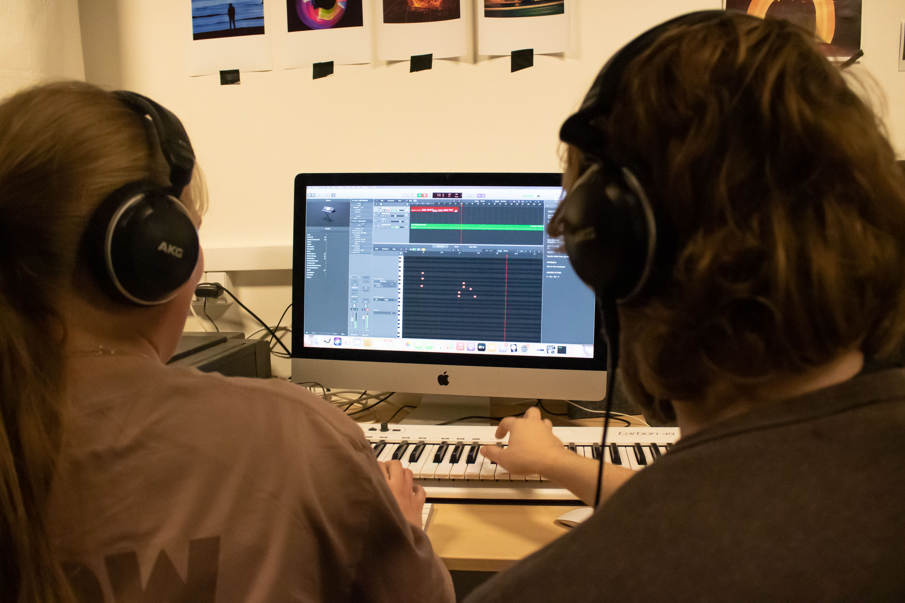 To personer med høretelefoner sidder omkring en iMac og producerer musik. Den ene spiller på et midi-keyboard.